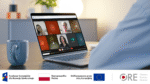 Osoba siedzącą przy biurku z laptopem. Na ekranie laptopa widać spotkanie online. Bierze w nim udział dziewięciu uczestników.