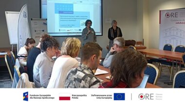 na pierwszym planie uczestnicy spotkania siedzący przy stołach, w tle ekran z wyświetloną prezentacją i tytułem projektu Opracowanie koncepcji i odbiór e-materiałów edukacyjnych ukierunkowanych na wspieranie kształcenia kompetencji zawodowych, przed ekranem dwie osoby, Joanna Saska-Dymnicka kierownik projektu oraz Anna Pregler główny ekspert merytoryczny projektu